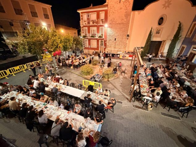 Imagen: Vista aérea nocturna de una plaza llena de gente cenando en mesas largas durante un evento popular en Parcent.
