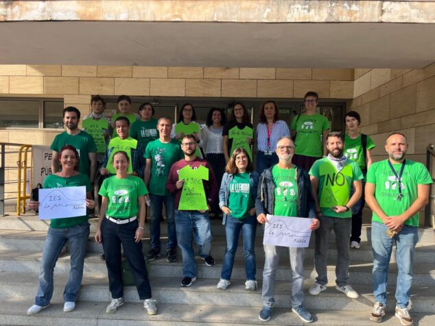 Grupo de profesores y profesoras posan en las escaleras de entrada del IES La Mar en Xàbia, muchos con camisetas verdes, sosteniendo carteles con lemas sobre la educación pública, ratios de alumnos y la lengua valenciana.