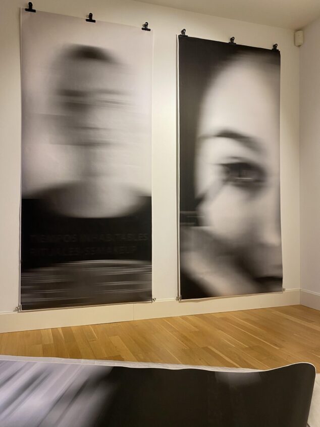 Imagen: Dos grandes pancartas fotográficas en blanco y negro con retratos borrosos colgando en el interior de una galería de arte con suelo de madera.