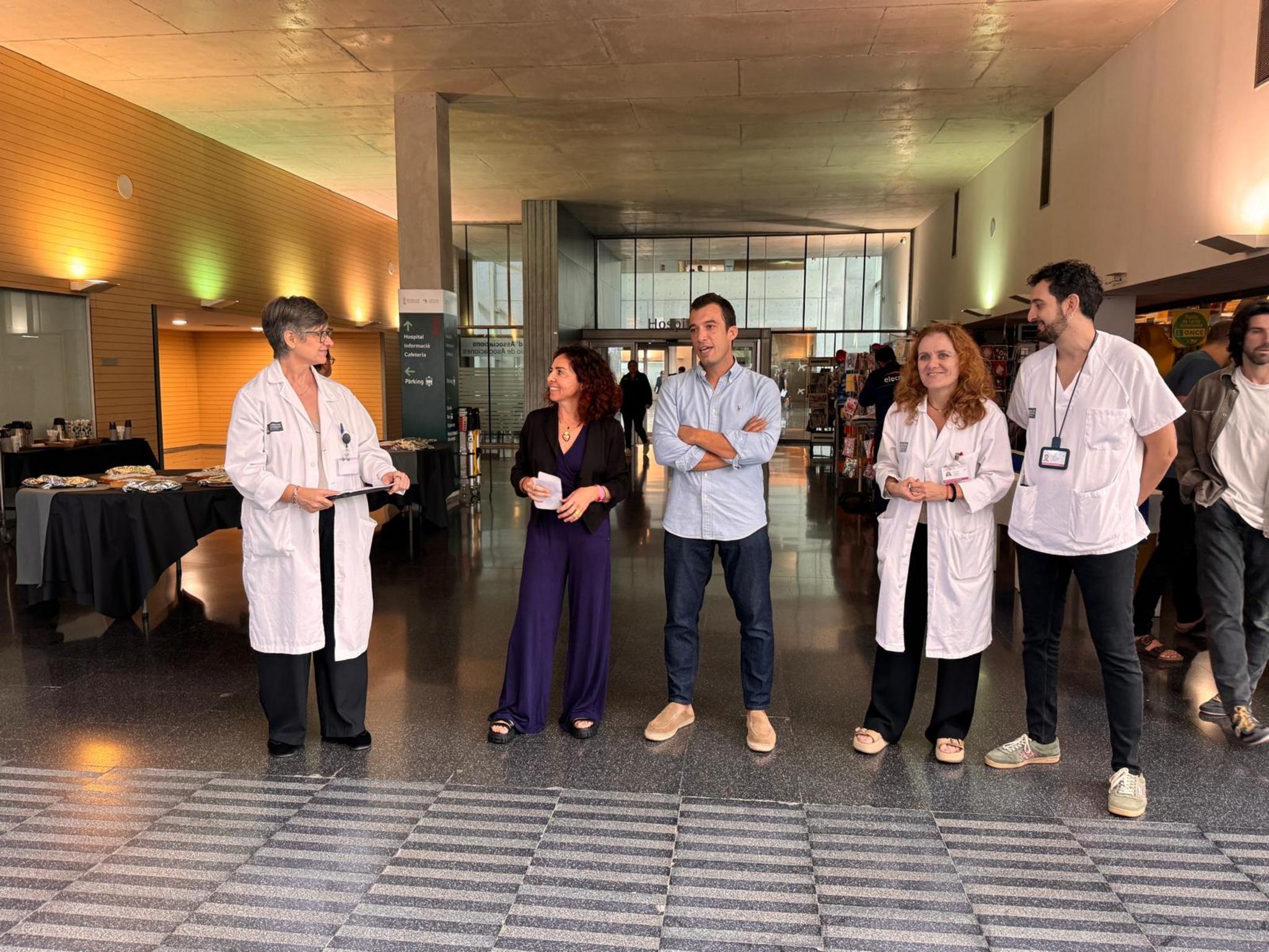 Varias personas, incluyendo profesionales con batas blancas, de pie en el vestíbulo del Hospital de Dénia junto a mesas informativas.