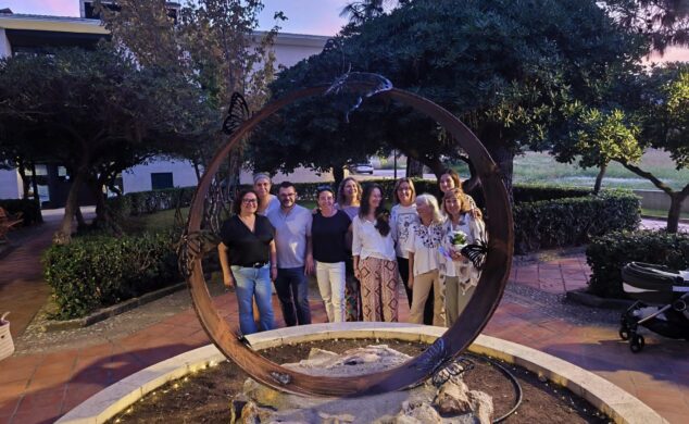 Imagen: Grupo de nueve personas posando de pie dentro y alrededor de una gran escultura circular de metal con mariposas en un parque al atardecer.