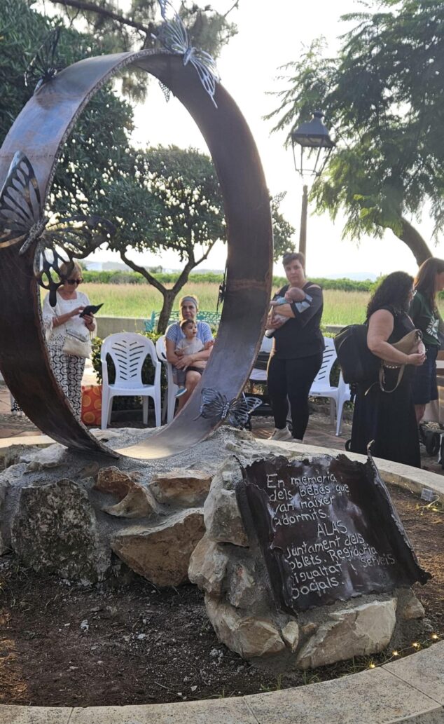 Imagen: Grupo de personas, incluyendo mujeres con niños pequeños, observan una escultura de metal con una placa conmemorativa en un parque.