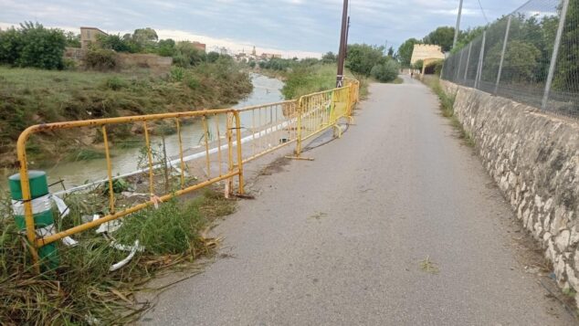 Imagen: Un camino asfaltado junto a un río crecido, con vallas amarillas de seguridad protegiendo una zona donde el margen se ha erosionado.