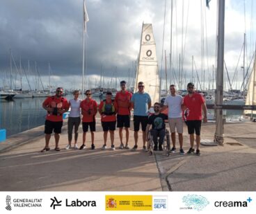 Grupo de personas, incluyendo instructores y participantes con chalecos salvavidas, posan en un muelle con veleros detrás durante la actividad Dia Blau en Dénia.