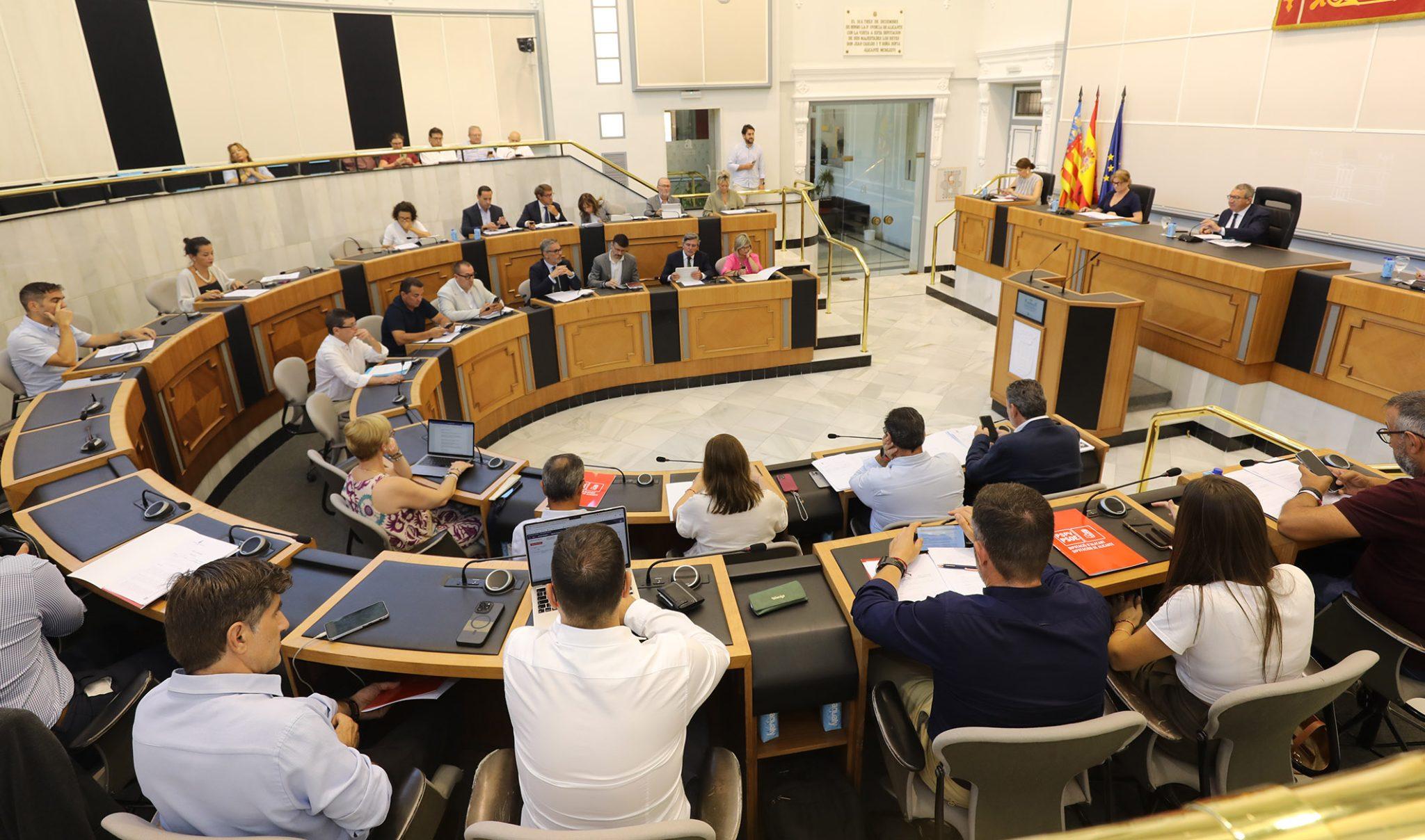 Fotografía de una sesión plenaria en el salón de la Diputación de Alicante, con políticos sentados en un hemiciclo y la mesa presidencial al fondo.