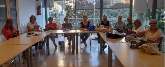 Varias mujeres sentadas alrededor de una mesa tejiendo ganchillo en un aula luminosa.